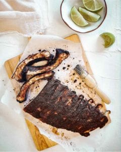 A rack of Vietnamese pork ribs on a sheet of parchment on a cutting board with a basting brush and a bowl of lime wedges nearby.