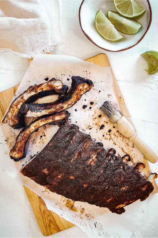 A rack of Vietnamese pork ribs on a sheet of parchment on a cutting board with a basting brush and a bowl of lime wedges nearby.