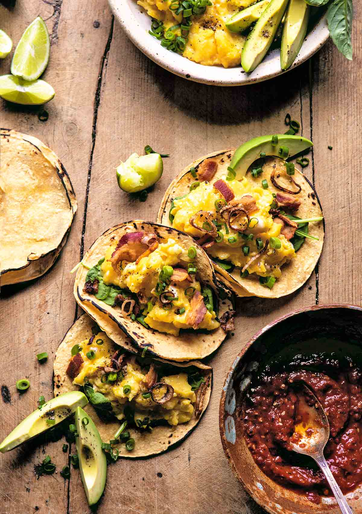Three avocado breakfast tacos with eggs, bacon, shallots, and chives with a bowl of salsa on the side.