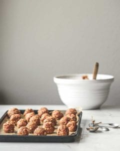 A tray of unbaked bbq meatballs with a set of measuring spoons beside it and a bowl of barbecue sauce in the background.