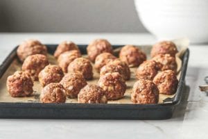 A tray of unbaked bbq meatballs with a set of measuring spoons beside it and a bowl of barbecue sauce in the background.