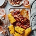 A tray of chicken thighs with balsamic barbecue sauce, corn on the cob, glasses of beer, forks, and plates