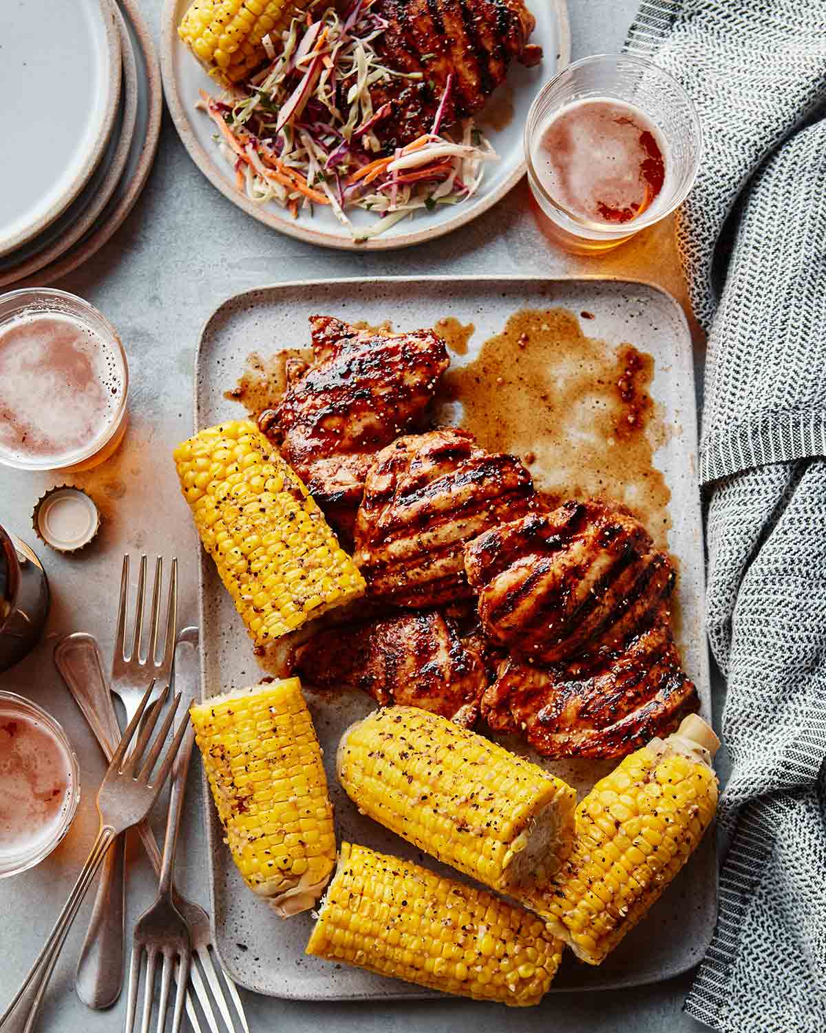 A tray of chicken thighs with balsamic barbecue sauce, corn on the cob, beer glasses, forks and plates