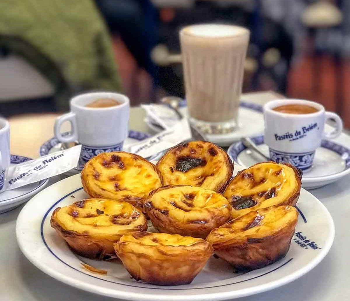 Seven pasteis de Belem, or Portuguese custard pastries, on a plate, with coffee cups nearby