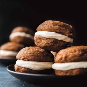 Two plates each with four pumpkin spice whoopie pies with cream cheese filling stacked on them.