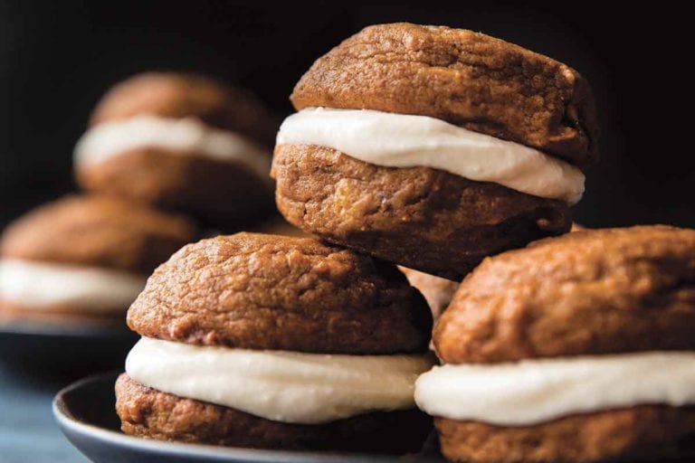 Two plates each with four pumpkin spice whoopie pies with cream cheese filling stacked on them.