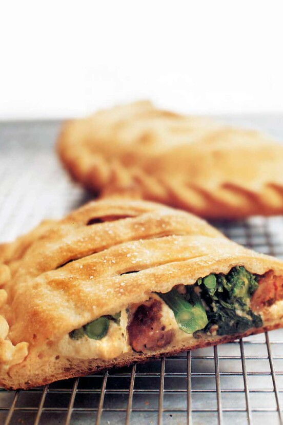 Two ricotta calzones with sausage and broccoli rabe, one cut in half, the other whole, on a wire rack.