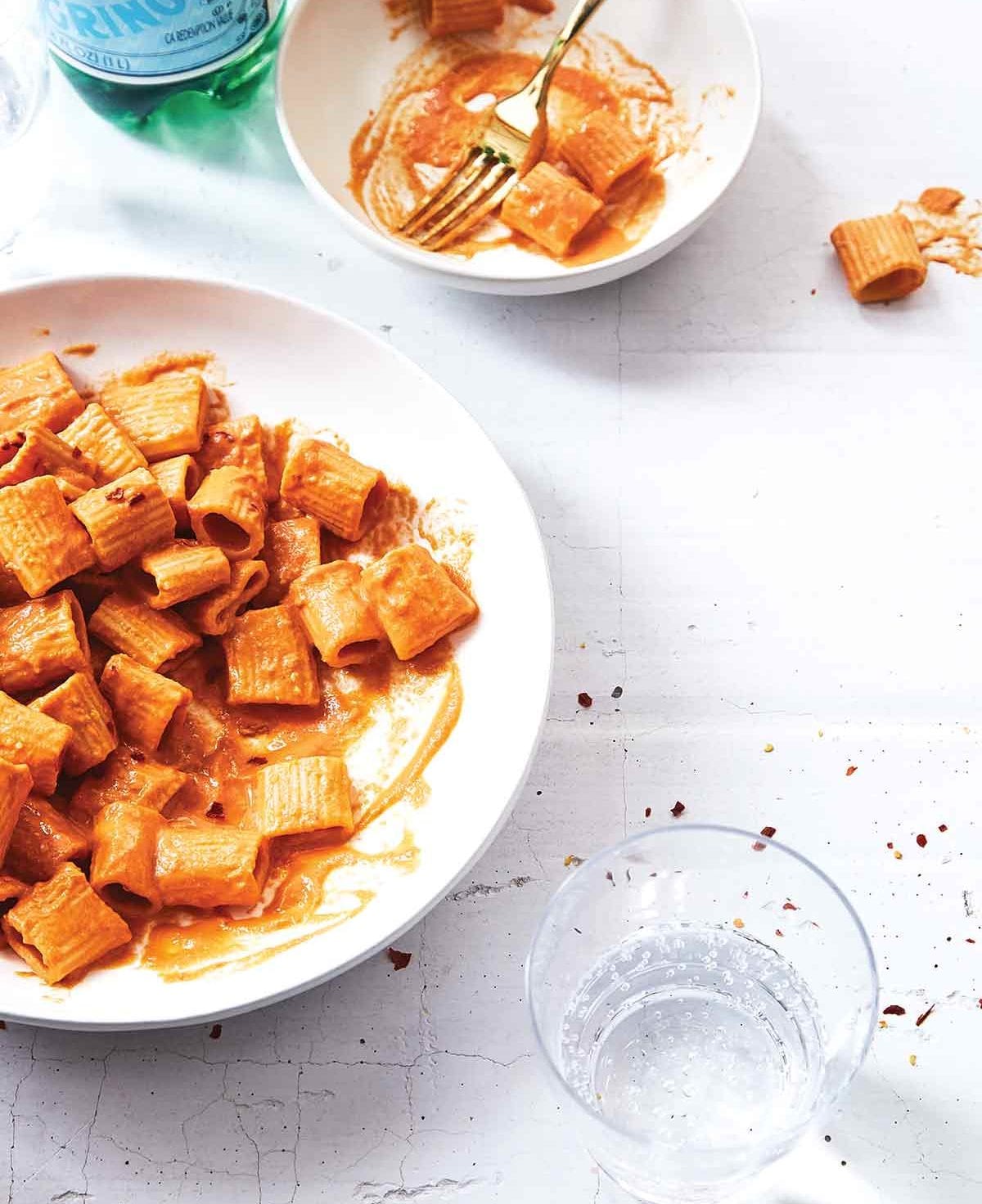 A white serving bowl and smaller white bowl partially filled with rigatoni with vegan vodka sauce and a glass of water on the side.