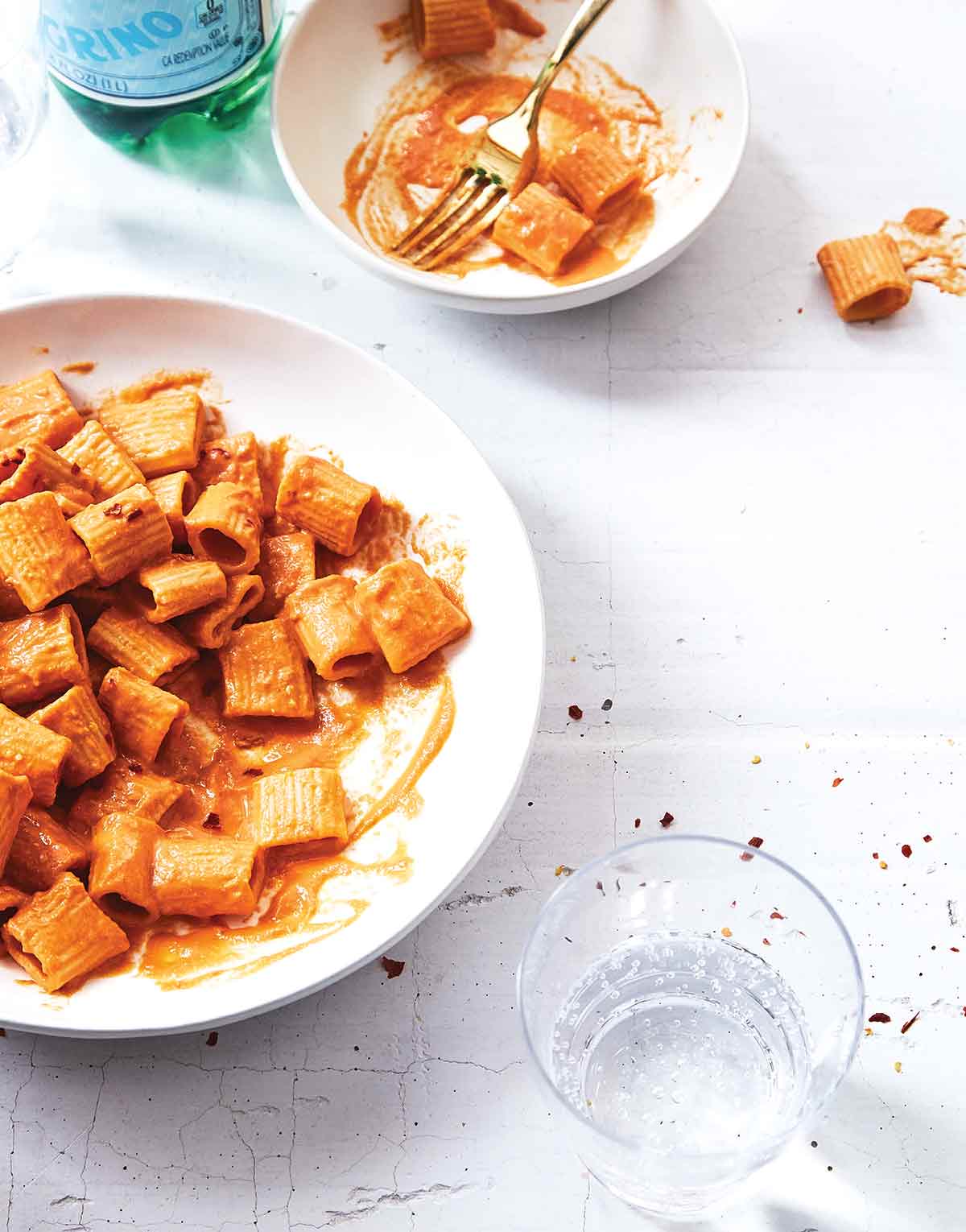 A white serving bowl and smaller white bowl partially filled with rigatoni with vegan vodka sauce and a glass of water on the side.