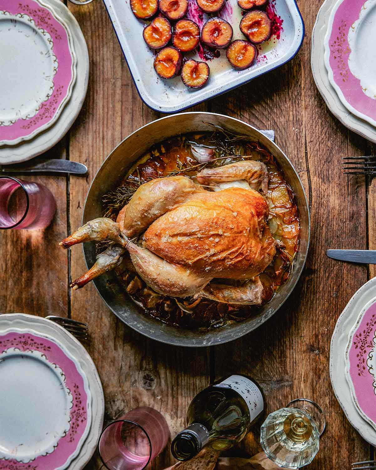 A whole roast chicken with rosemary and potatoes on a wooden table with four plates and a tray of roasted plums beside it.