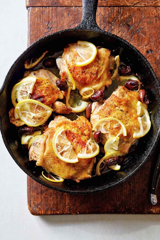 Four roast chicken thighs with lemon in a cast-iron skillet set on a wooden board with a meat fork resting beside it.