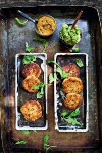 Six salmon cakes on rectangular platters with bowls of avocado spread and chimichurri beside.