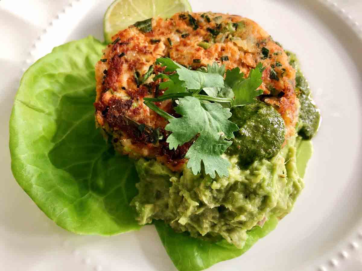 A salmon cake with a scoop of avocado spread, a drizzle of chimichurri, and a garnish of cilantro on a butter lettuce leaf.
