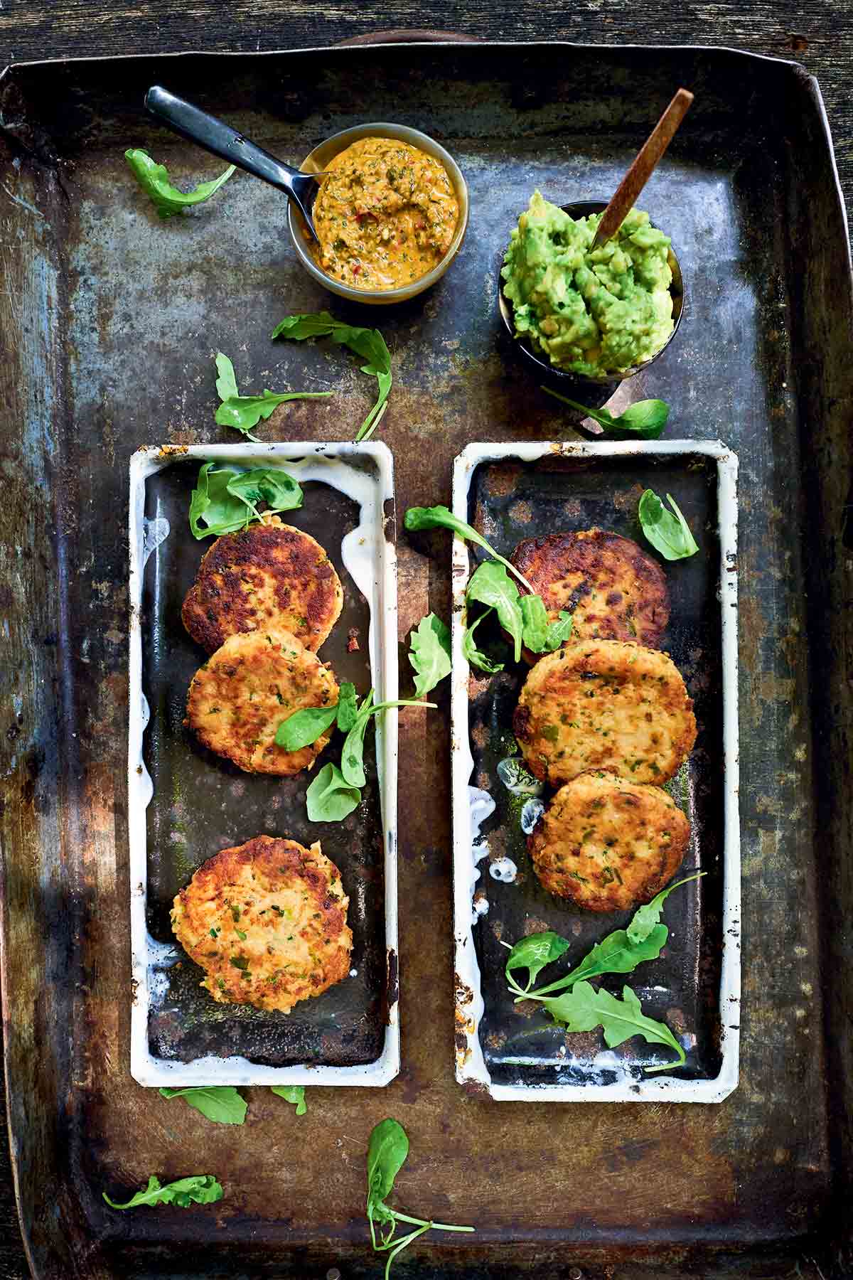 Six salmon cakes on rectangular platters with bowls of avocado spread and chimichurri beside. Perfect for sustainability.