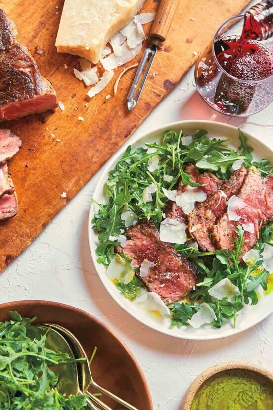 A steak and arugula salad with pesto vinaigrette in a white bowl, with a wooden board and sliced steak next to it.