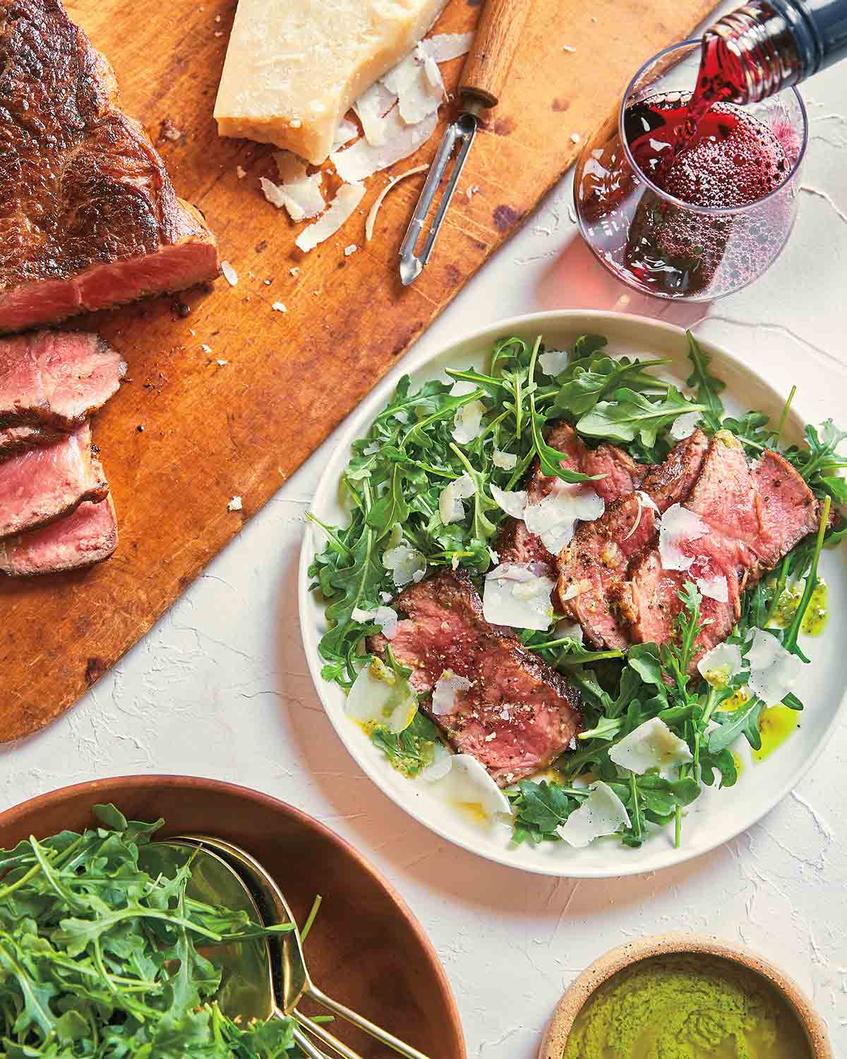 A steak and arugula salad with pesto vinaigrette in a white bowl, with a wooden board and sliced steak next to it.