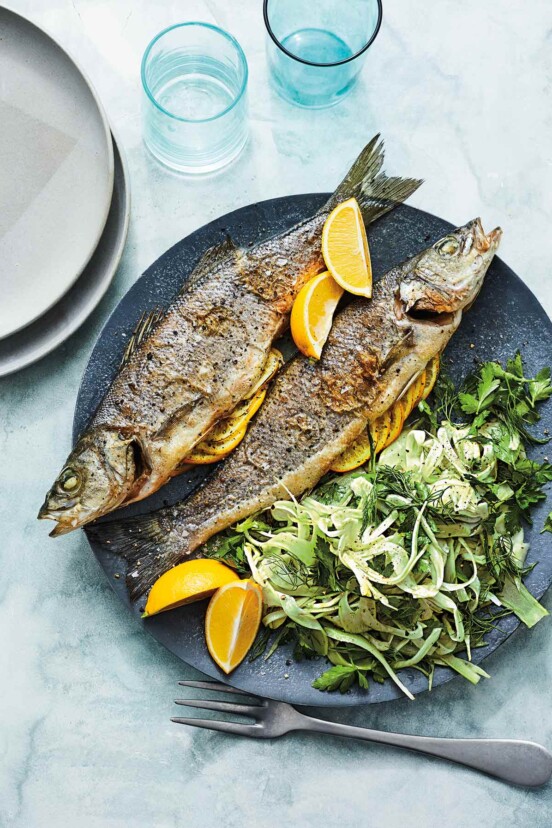 Two whole roasted branzino with lemon and shaved fennel salad on a blue plate with a fork, two glasses, and two grey plates beside it.