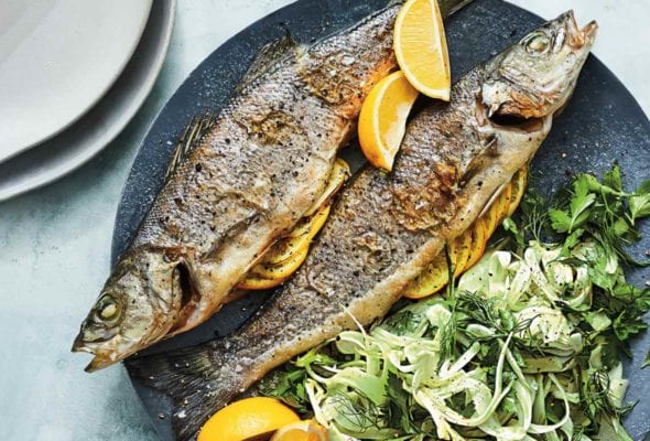 Two whole roasted branzino with lemon and shaved fennel salad on a blue plate with a fork, two glasses, and two grey plates beside it.