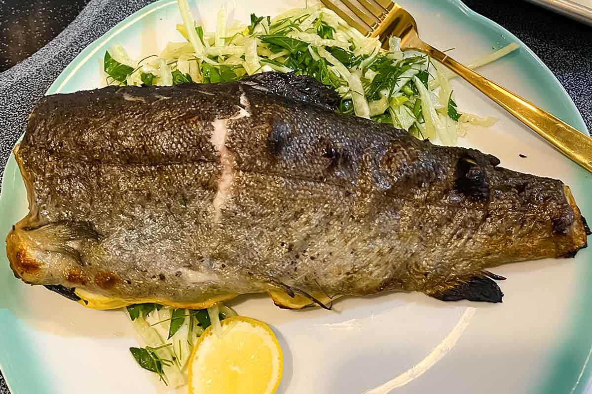 A whole roasted branzino with lemon and shaved fennel salad on a blue and white plate with a gold fork resting beside the salad.