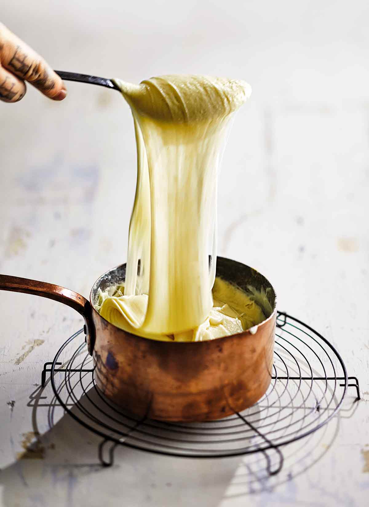 A person holding a spoonful of aligot potatoes over a copper pot.