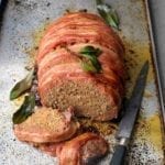 A partially sliced bacon-wrapped meatloaf on a rimmed baking sheet with a knife beside it and bay leaves scattered around it.