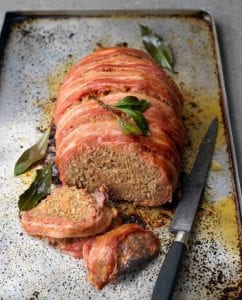 A partially sliced bacon-wrapped meatloaf on a rimmed baking sheet with a knife beside it and bay leaves scattered around it.