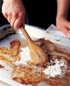 A person stirring flour into the basic pan gravy with a wooden spoon on a rimmed baking sheet.