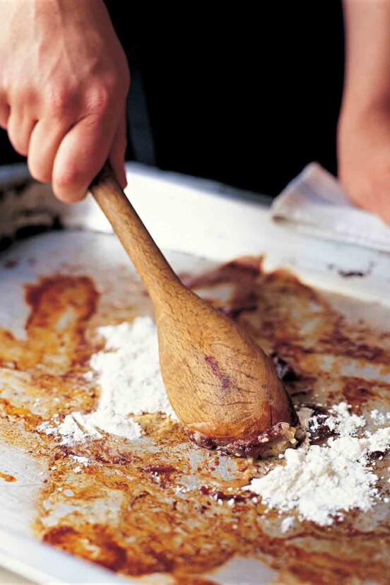 A person stirring flour into the basic pan gravy with a wooden spoon on a rimmed baking sheet.