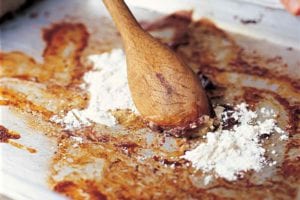 A person stirring flour into the basic pan gravy with a wooden spoon on a rimmed baking sheet.