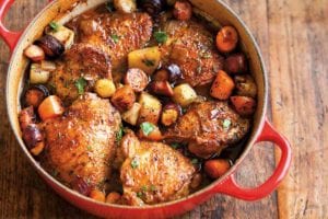 A red Dutch oven filled with beer-braised chicken with root vegetables on a wooden surface.