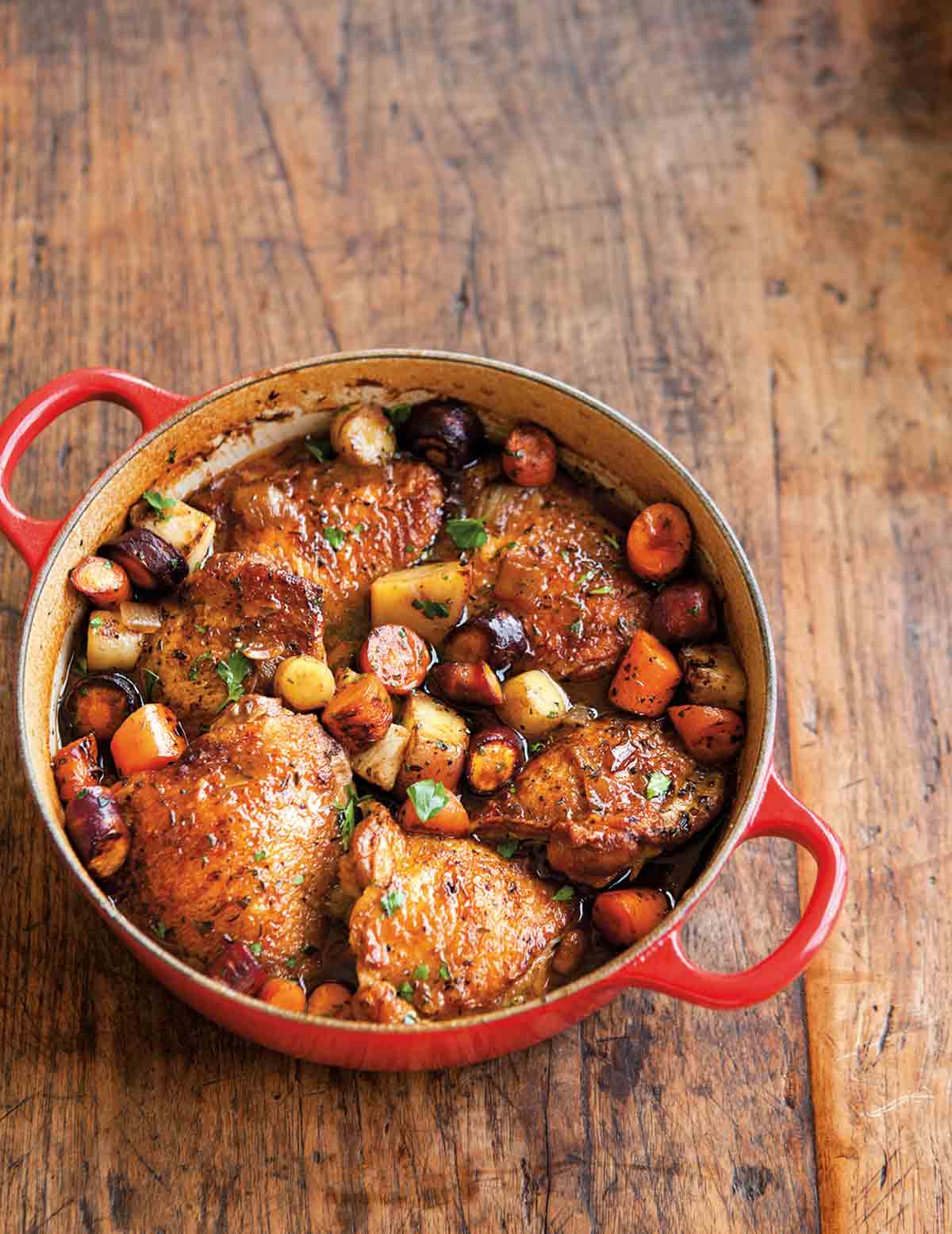 A red Dutch oven filled with beer-braised chicken with root vegetables on a wooden surface.