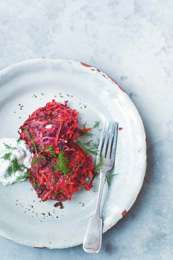Two beet and carrot fritters, garnished with dill, on a white plate with a fork and a dollop of dill yogurt