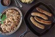 Four cooked bratwurst and sauerkraut, along with mashed potatoes and grainy mustard in separate dishes beside the skillet containing the bratwurst.