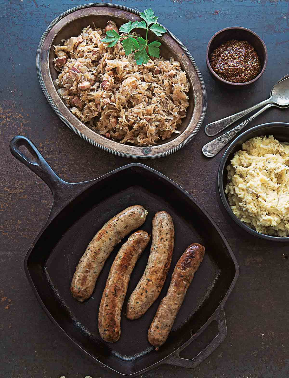 Four cooked bratwurst and sauerkraut, along with mashed potatoes and grainy mustard in separate dishes beside the skillet containing the bratwurst.