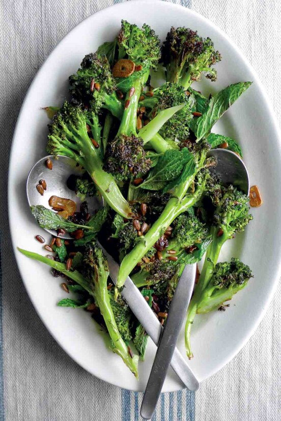 A white plate with spears of roasted broccoli with pickled golden raisins scattered overtop and two spoons in the broccoli.