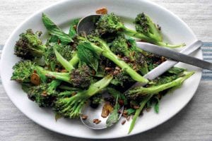 A white plate with spears of roasted broccoli with pickled golden raisins scattered overtop and two spoons in the broccoli.