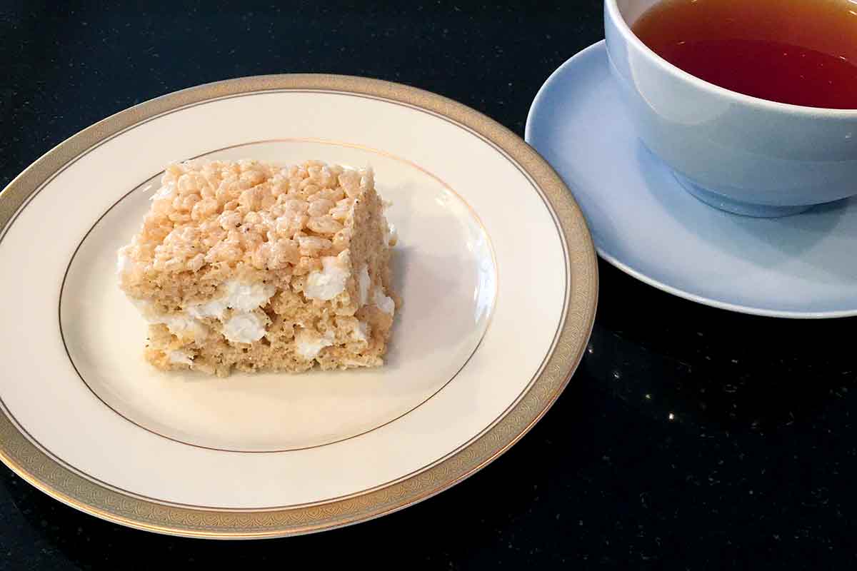 A brown butter rice Krispies treat on a china plate next to a cup of tea.