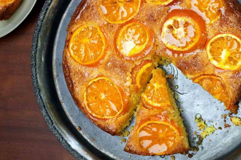 A clementine cake on a round silver platter with one slice on a plate beside it.