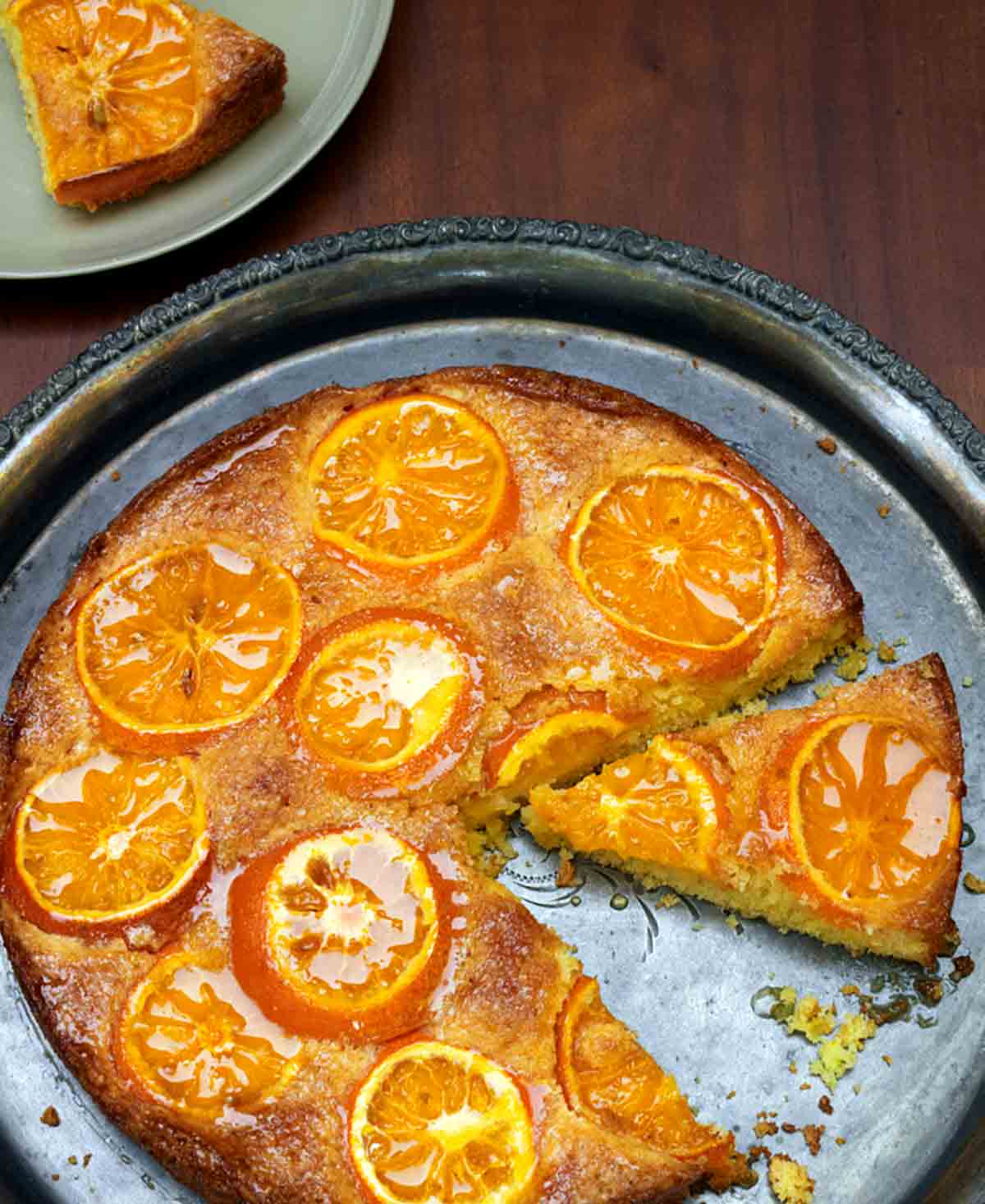 A clementine cake on a round silver platter with one slice on a plate beside it.