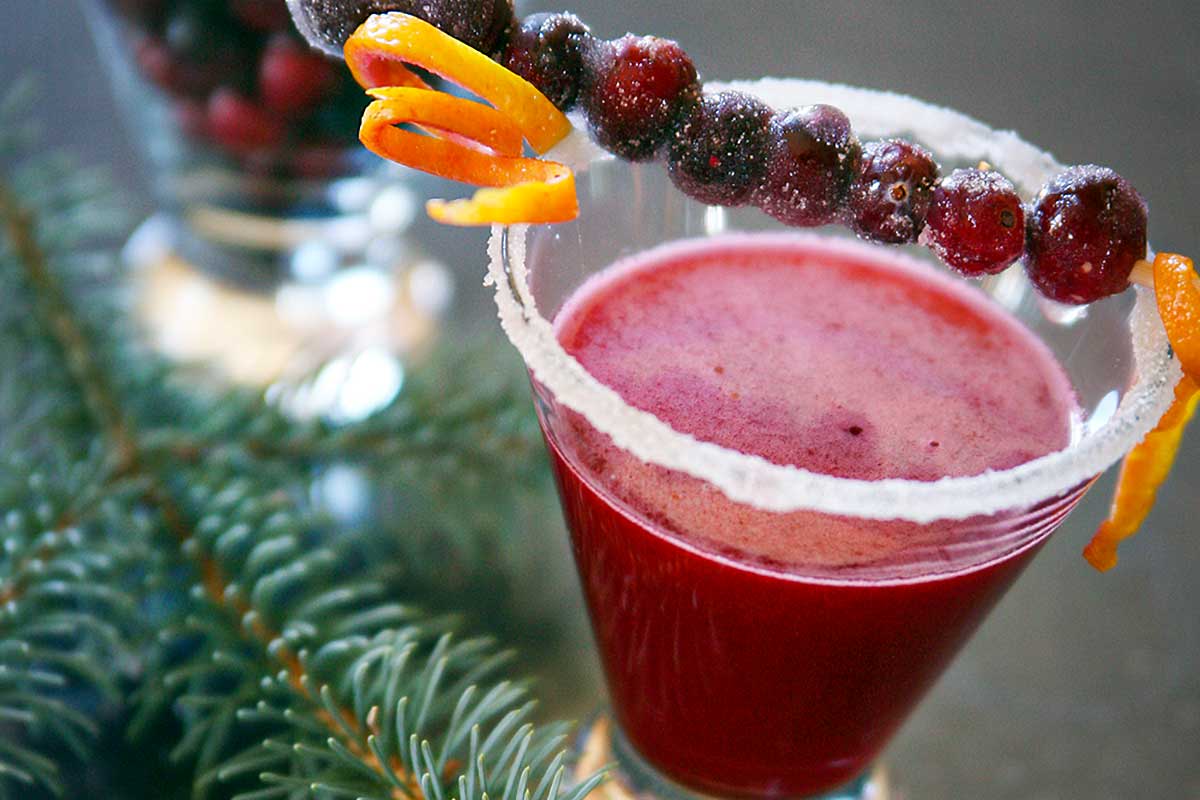 A cranberry margarita in a short glass with a skewer or sugared cranberries and orange zest across the top.