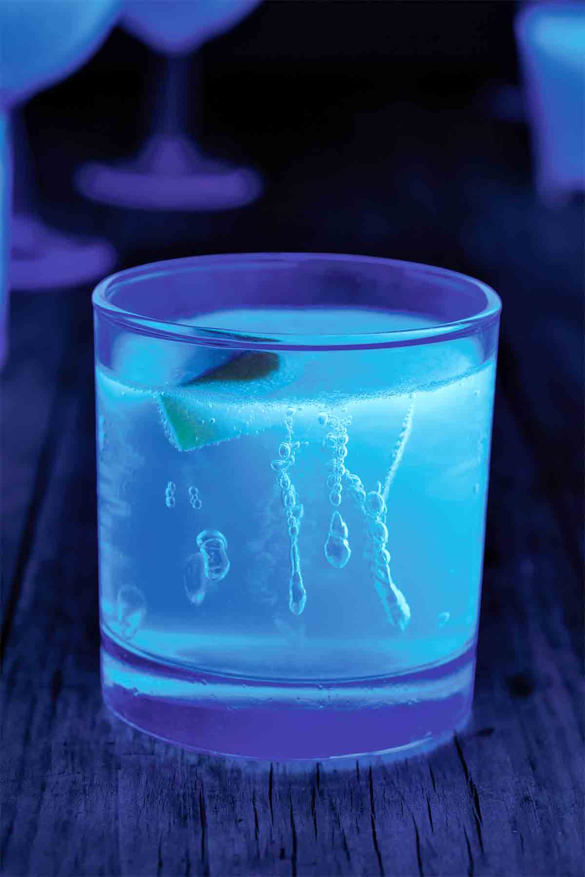 A rocks glass filled with neon blue glow-in-the-dark Jello on a dark background.