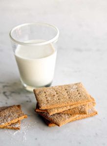 A pile of homemade graham crackers on a marble table with a glass of milk nearby.