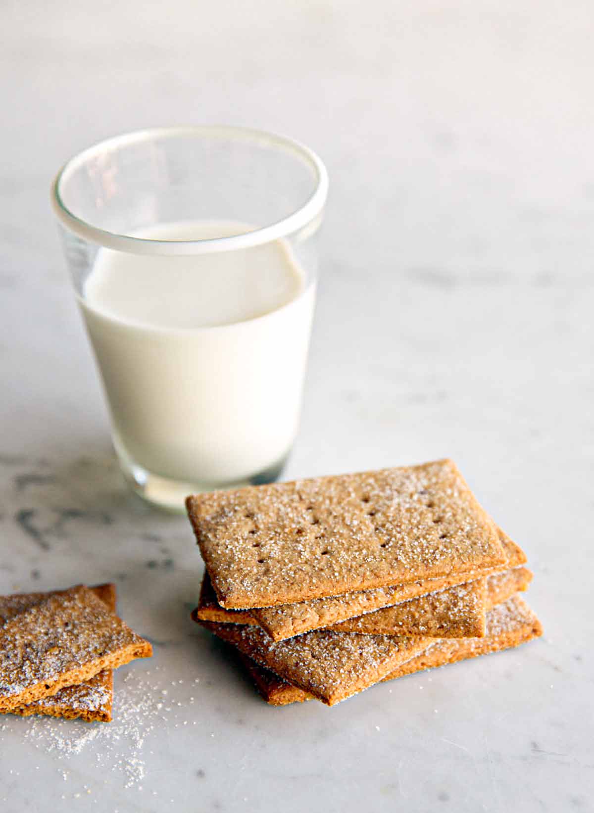 A pile of homemade graham crackers on a marble table with a glass of milk nearby