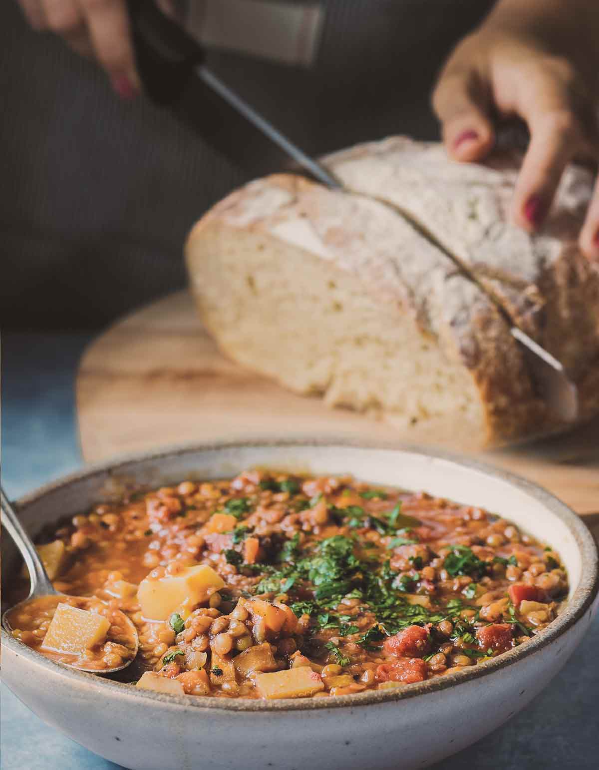 A bowl of Instant Pot lentil soup, with a person cutting a slice of artisan bread in the background.