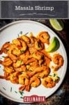 A white plate filled with masala shrimp, with lime wedges on the side, and gives sprinkled over, to garnish.