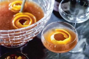 A glass punch bowl partially filled with maybelle punch and orange peel garnish, and two coupe glasses alongside, one filled and one empty.