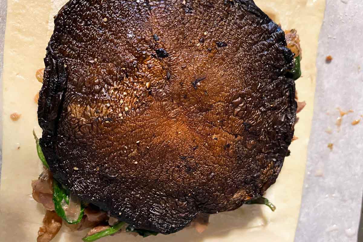 A roasted portobello mushroom on top of filling and pastry in the preparation of mushroom wellingtons with spinach and walnuts.