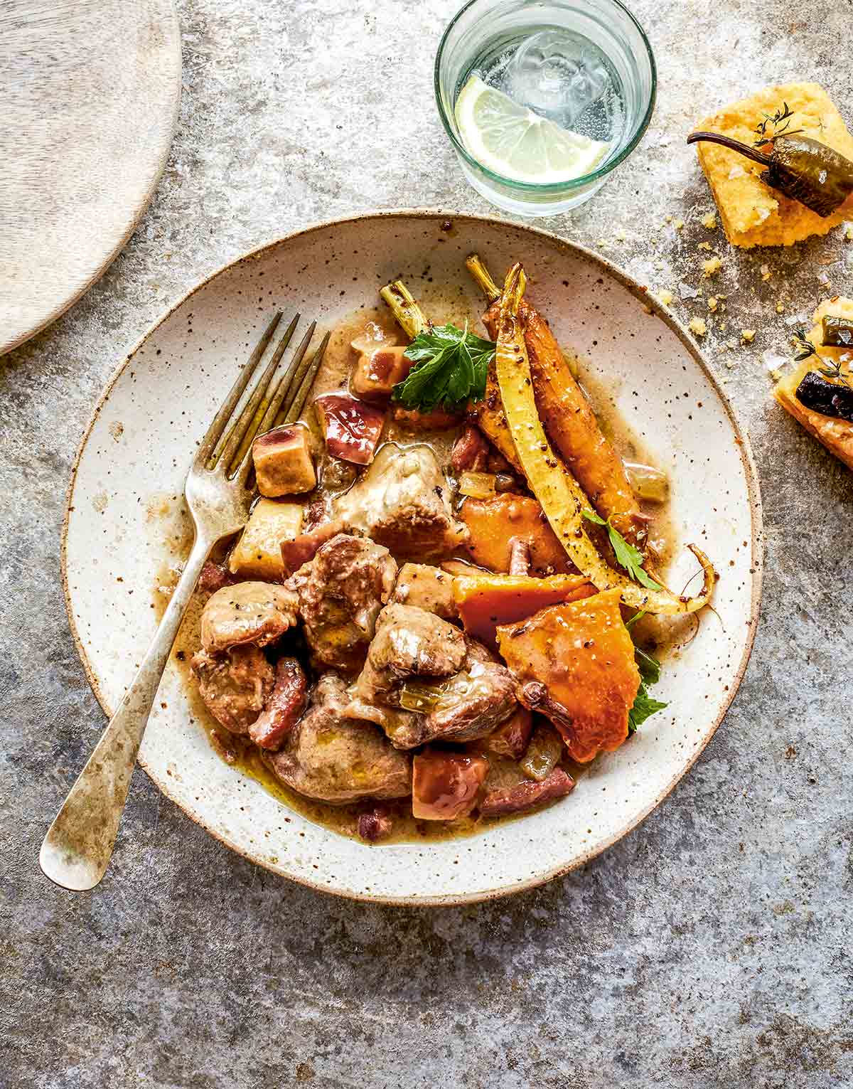 A white speckled bowl filled with pork casserole with apples and cider, roasted carrots, and a fork resting on the side.