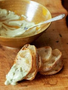 A bowl with creamy green olive dip behind five slices of baguette with dip on one