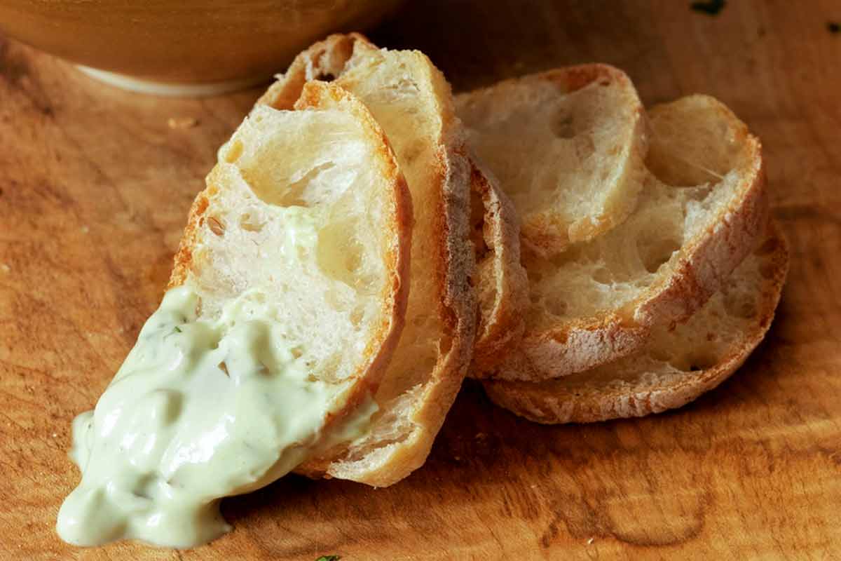 A bowl with creamy green olive dip behind five slices of baguette with dip on one